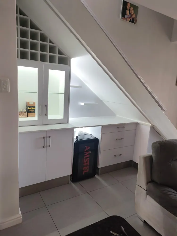 A bar built into the space under a staircase, with white gloss cupboards, a wine rack fitted to the rake of the stairs and lit glass-fronted cabinets.